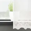 A close-up view of a white lace curtain with a floral pattern, partially obscuring a potted plant on a windowsill. Produktbild-Vorschau