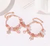 Two rose gold charm bracelets with pink and clear beads and charms are displayed on a white surface with a blurred white object and a card with text in the background. Produktbild-Vorschau