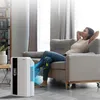 A woman relaxing on a couch while an air purifier operates in the foreground. Náhled obrázku produktu