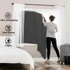 A woman is hanging a dark gray blackout fabric in front of a window, with white sheer curtains on either side. A bed is visible on the left, and a suitcase on the right. Produktbild-Vorschau