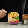 A person is using a fork to eat from a glass lunch container filled with layered food. Podgląd zdjęcia produktu