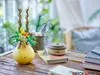 A LEGO flower pot with a bouquet of flowers sits on a wooden table, with books and a spool of twine nearby. The background shows a blurred indoor setting with greenery and furniture. Podgląd zdjęcia produktu