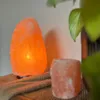 A close-up shot of two Himalayan salt lamps, one larger and lit, and a smaller one next to it, placed on a textured fabric surface. Produktbild-Vorschau