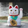 A white Maneki-neko cat figurine sits on a textured surface with a blurred background of water and greenery. Produktbild-Vorschau