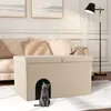 A cat sits in front of a beige upholstered storage ottoman with a cat-sized entrance, placed in a living room setting. Produktbild-Vorschau