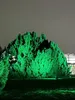 A large, lush green tree illuminated by green lights at night, with a greenhouse structure visible in the background. Produktbild-Vorschau