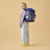 A young girl wearing a backpack stands against a plain background. Produktbild-Vorschau