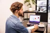 A man wearing earbuds is working on a laptop in a cafe. Produktbild-Vorschau