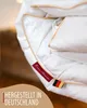 A close-up of a white duvet with gold trim, showing brand tags and a German flag. Produktbild-Vorschau
