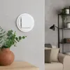 A close-up of a white digital thermostat mounted on a textured gray wall, with a terracotta vase containing eucalyptus leaves in the foreground and a blurred living room scene in the background. Anteprima dell’immagine prodotto