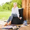 A woman is sitting on a large black beanbag chair outdoors on a patio, with a colorful rug in front of her and a pitcher and glass nearby. Lush green trees and a wooden wall form the background. Produktbild-Vorschau