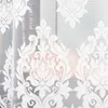 A close-up view of a white lace curtain with a damask pattern, partially obscuring a person in the background. Náhled obrázku produktu