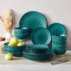 A collection of teal-colored ceramic dinnerware with a spiral pattern, including plates and bowls, arranged on a light-colored surface with some pears and dried grass in a vase. Produktbild-Vorschau