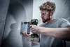A man is using a cordless impact driver to attach a metal bracket to a concrete wall. Produktbild-Vorschau