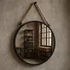 A round mirror with a rope hanger is mounted on a textured wall. The mirror reflects a room with industrial-style furniture, including a tall shelf unit and a console table. Produktbild-Vorschau
