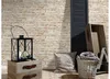 A decorative vignette with a stone-effect wallpaper background, featuring a lantern on a wooden chest, a pillow, and a basket with blankets. Produktbild-Vorschau