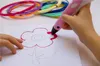 A child's hands are using a pink 3D pen to draw a flower on a piece of paper. Several spools of colorful filament are visible in the background. Náhľad obrázku produktu