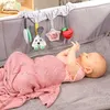 A baby lying on a bed covered with a pink blanket, reaching for toys hanging from a crib. Produktbild-Vorschau