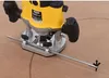 A close-up of a yellow and black router tool with a metal guide rod attached, resting on a brown surface with faint circular markings. Náhled obrázku produktu
