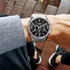 A person wearing a silver and black Casio watch on their wrist, with a grey shirt and black shoes visible. Produktbild-Vorschau