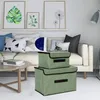 Two green storage boxes stacked on a couch next to decorative pillows, with a coffee table and artwork in the background. Produktbild-Vorschau