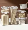A collection of clear plastic food storage containers filled with various dry goods, arranged on a white surface against a brown background. Produktbild-Vorschau
