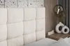 A close-up view of a bed with a tufted headboard, a bedside table with decorative vases and a small 'home' sign, and a hanging lamp. Produktbild-Vorschau