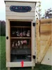 A wooden smoker with meat hanging inside, set up outdoors on grass. Náhled obrázku produktu