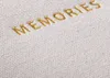 A close-up of the word "MEMORIES" embroidered in gold thread on a textured white fabric. Podgląd zdjęcia produktu