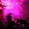 A room is illuminated by a pink and green star projector, creating a galaxy effect on the walls and ceiling. Plants and furniture are visible in the dimly lit space. Podgląd zdjęcia produktu
