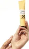 A hand holds a tube of sunscreen against a white background. Produktbild-Vorschau