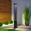A modern black bollard light stands on a wooden deck in a garden setting, illuminated by its own light. Anteprima dell’immagine prodotto