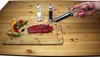 A hand is using a stainless steel marinade injector on a raw steak on a wooden cutting board. Other ingredients like chili peppers, rosemary, and peppercorns are also on the board. Produktbild-Vorschau