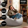 A young woman is sitting on a large, round, gray beanbag chair in a bedroom, listening to music with headphones. Produktbild-Vorschau