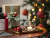 A festive Christmas scene with gifts, a lit candle in a glass holder, and a decorated Christmas tree in the background. Produktbild-Vorschau