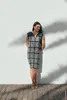 A woman stands against a white wall, leaning slightly, with shadows of leaves cast on the wall and ground. Produktbild-Vorschau