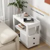 A white side table with drawers and shelves is placed next to a cream-colored sofa. A vase with greenery, a decorative bowl, and a cup of coffee on a book are on the top shelf. Produktbild-Vorschau