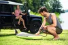A man is inflating a paddleboard next to a car, with a woman sitting in the car. Produktbild-Vorschau