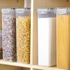 Four clear plastic food storage containers with white lids are arranged on a shelf. The containers are filled with different dry goods: the first with what appears to be rice or grains, the second with fusilli pasta, the third with flour, and the fourth wi Produktbild-Vorschau