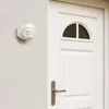 A white motion sensor is mounted on a textured white wall next to a white door with a semi-circular window and a silver door handle. Produktbild-Vorschau