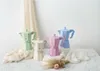 Four colorful Moka pots are arranged on a marble surface with draped fabric and dried flowers. Anteprima dell’immagine prodotto