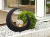 A decorative solar lamp in a crescent shape with a glowing sphere inside, placed in a garden setting with gravel and a bush. Anteprima dell’immagine prodotto