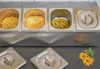 A stainless steel food topping dispenser with various ingredients inside, placed on a dark surface with some decorative flowers. Produktbild-Vorschau
