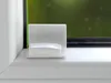 A close-up view of a white plastic mounting bracket with a green cord attached, placed on a white windowsill next to a window frame. Produktbild-Vorschau