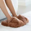 A person's legs and feet are shown wearing plush brown slippers while resting on a white rug. Náhľad obrázku produktu