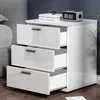A white nightstand with three drawers is shown in a bedroom setting, next to a gray upholstered headboard. Podgląd zdjęcia produktu