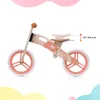 A wooden balance bike with a pink and orange abstract pattern, shown in profile against a white background with soft pastel color blobs at the bottom and top. Náhľad obrázku produktu
