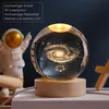 A crystal ball with a galaxy engraving sits on a wooden base, with an astronaut figurine in the background. Produktbild-Vorschau