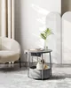 A cat is sitting in a pet bed integrated into a round side table with built-in power outlets and USB ports. The table has a plant in a vase on top, and a phone is charging. A chair is visible in the background. Produktbild-Vorschau