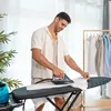 A man is ironing a white shirt on an ironing board in a well-lit room with clothes hanging on a rack in the background. Náhled obrázku produktu